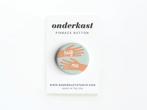The Hug Me Hands Pinback Button by Onderkast Studio features a hand gesture and hug me text, displayed on a card labeled Onderkast Studio PINBACK BUTTON with the website and MADE IN THE USA below.