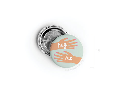 The Hug Me Hands Pinback Button by Onderkast Studio features two illustrated arms reaching to hug with “hug me” in white on light green. Printed in the USA, this round button measures 1.25 in diameter.