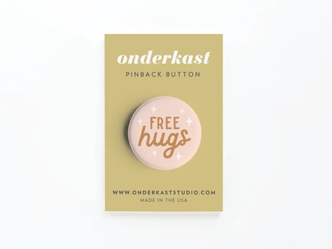 The Free Hugs Pinback Button by Onderkast Studio features white stars on beige, attached to a tan branded card. It’s made in the USA, with a sturdy metal pin and website info printed at the bottom.