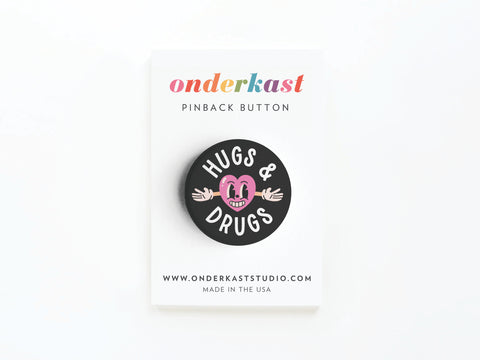 The Hugs & Drugs Pinback Button by Onderkast Studio is a round black button with a pink smiling cartoon figure and vibrant colors, representing playful touch, displayed on a white Onderkast pinback button card.