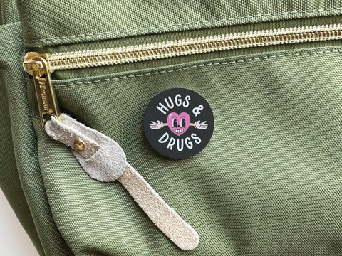 A round black Hugs & Drugs Pinback Button by Onderkast Studio, with a smiling pink cartoon face and outstretched arms on vibrant colors, features “HUGS & DRUGS” in white encircling the playful design.