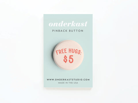 The Free Hugs $5 Pinback Button from Onderkast Studio features bold red text and comes on a light blue branded card with the website and MADE IN THE USA printed at the bottom.