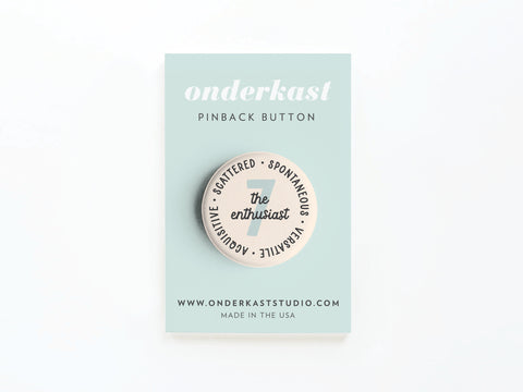 A light blue Onderkast Studio card displays the Enneagram Pinback Button – 7 The Enthusiast, featuring words like SCATTERED, SPONTANEOUS, VERSATILE, and ADOPTIVE—a fun personality type accessory.