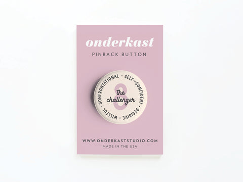 The Enneagram Pinback Button – 8 The Challenger by Onderkast Studio is round and displayed on a pink card, featuring “Confrontational, Self-confident, Decisive, Willful” around the edge.