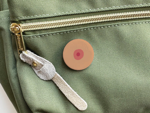The Onderkast Studio Boob Pinback Button – Medium, featuring an abstract nipple design, is attached to the front pocket of a light green bag with a gold zipper and suede pull to celebrate self-love and body positivity.