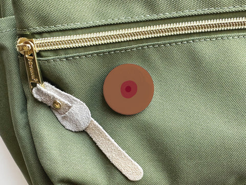 The Boob Pinback Button – Dark by Onderkast Studio, shaped like a brown nipple with a dark areola, is shown on a green bag near a gold zipper with suede pull—celebrating self-love and body positivity.