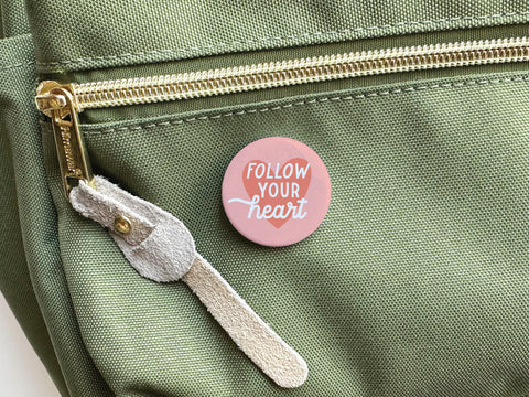 The Onderkast Studio Follow Your Heart Deep Pinback Button in pink adds whimsy when pinned to a light green bag featuring a gold zipper and suede pull.