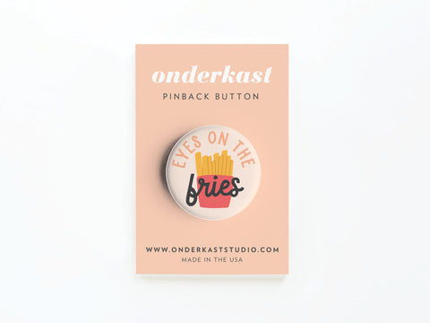 The Eyes on the Fries Pinback Button by Onderkast Studio features a vibrant fry design with EYES ON THE fries text, mounted on a peach card that notes onderkast PINBACK BUTTON, includes the website, and states MADE IN THE USA.