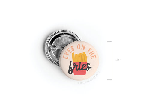 The Eyes on the Fries Pinback Button by Onderkast Studio features “EYES ON THE fries” above a red carton of yellow fries. At 1.25 inches, it’s a bold, fun accessory to show your love of snacks and style!.