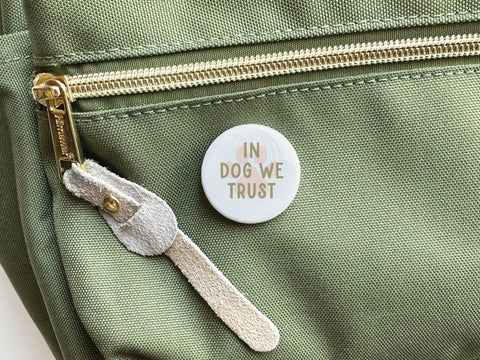 The Onderkast Studio In Dog We Trust Pinback Button, featuring light brown and orange text, is displayed on a green fabric bag with a gold zipper and beige suede pull tab—a perfect celebration of canine loyalty.