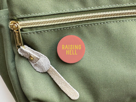 The Onderkast Studio Raising Hell Pinback Button—a round red pin with bold yellow text—adds rebellious flair when clipped to the pocket of a light green bag with a gold zipper and beige suede pull.