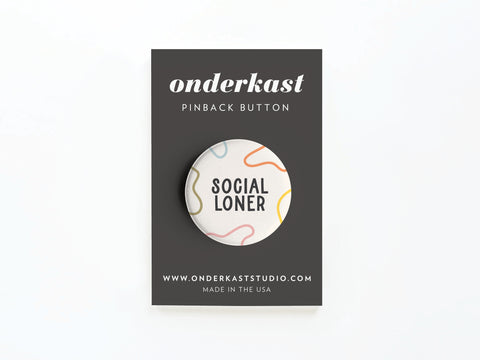 The Social Loner Pinback Button by Onderkast Studio features black text on white with colorful abstract squiggles, displayed on a dark gray onderkast card—a playful ode to independence and solitude.