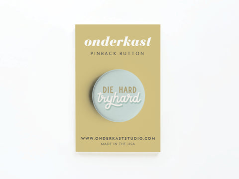 The Die Hard Tryhard Pinback Button by Onderkast Studio is light blue with bold white text, mounted on a tan card that reads onderkast pinback button. Card bottom shows website and Made in the USA.