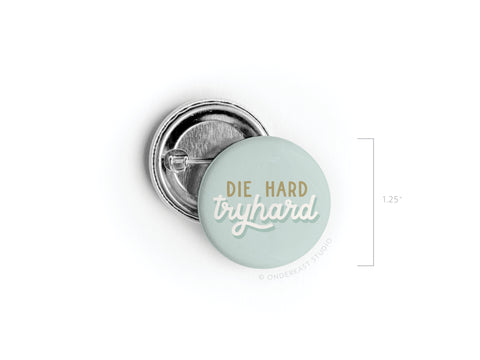 The Die Hard Tryhard Pinback Button by Onderkast Studio features a pale blue-green background with gold and white lettering. Shown with its metal pin-back and a 1.25-inch marker, this accessory makes a standout addition to any collection.