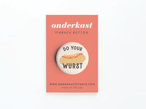 The Do Your Wurst Pinback Button by Onderkast Studio features a vibrant hot dog illustration and the text DO YOUR WURST, displayed on a coral card with onderkaststudio.com printed at the bottom.