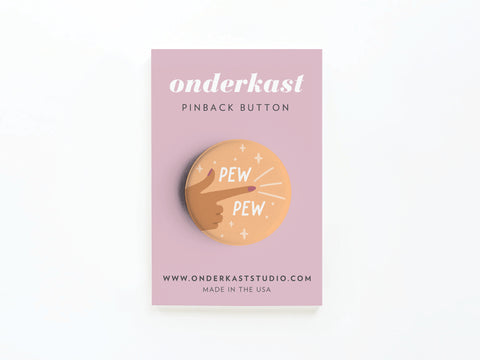 The Pew Pew Pinback Button by Onderkast Studio features a finger-gun hand with “PEW PEW” and sparkles on a pink card that says “Onderkast PINBACK BUTTON,” includes the website, and states MADE IN THE USA.