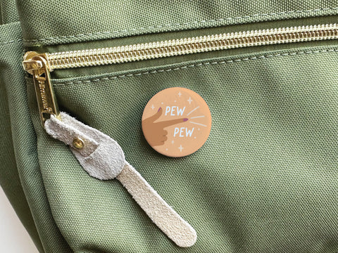 The Pew Pew Pinback Button by Onderkast Studio, featuring a hand making a finger-gun gesture and PEW PEW! with sparkles, is shown attached to a green fabric bag near a zippered pocket.