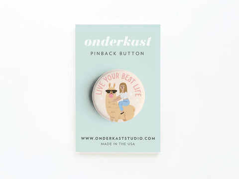 The Live Your Best Life Pinback Button by Onderkast Studio shows a girl riding a smiling llama in sunglasses with the phrase above. High-quality, its mounted on a blue card featuring onderkast pinback button, website, and Made in the USA.