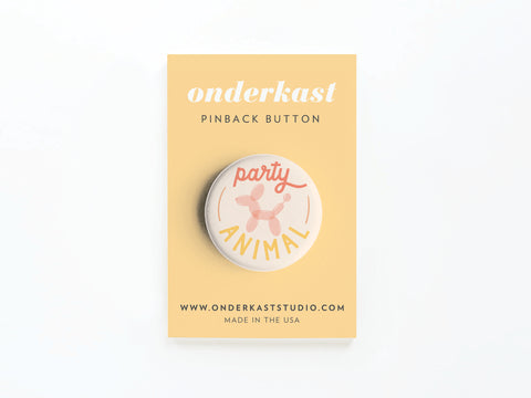 The Party Animal Pinback Button by Onderkast Studio features a vibrant pink balloon animal on a yellow card that says onderkast PINBACK BUTTON and includes www.onderkaststudio.com at the bottom.