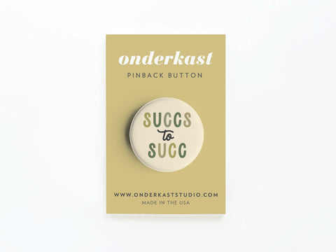 The Succs to Succ Pinback Button by Onderkast Studio features green and gray text on a button displayed on a beige card reading onderkast PINBACK BUTTON, with the website and MADE IN THE USA at the bottom.