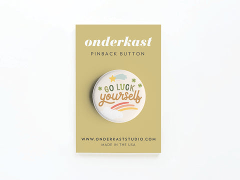 The Go Luck Yourself Pinback Button by Onderkast Studio features playful lettering with stars, clovers, and a rainbow. It’s attached to a tan card labeled Onderkast pinback button and is proudly printed in the USA.