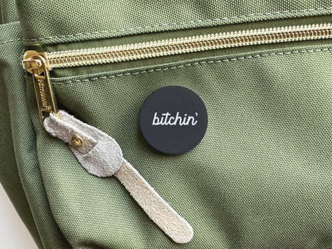 The Bitchin’ Pinback Button by Onderkast Studio, featuring bold white script, is attached to a light green bag with a gold zipper and suede pull. Its vibrant colors ensure this retro accessory stands out.