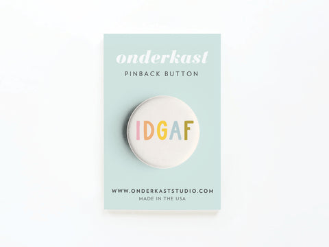 The Onderkast Studio IDGAF Pinback Button is a round, pastel accessory attached to a light blue card labeled with the brand and website, plus Made in the USA at the bottom.