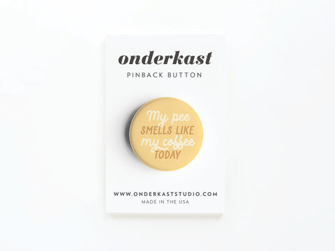 The discontinued “My Pee Smells Like Coffee Today” round pinback button by Onderkast Studio features vivid yellow and white text on a white card displaying the brand name and website details.