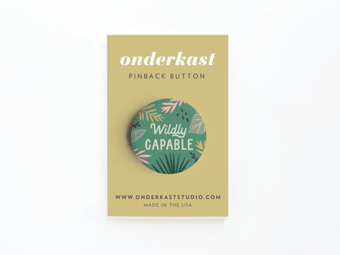 The Wildly Capable Pinback Button by Onderkast Studio features green leaves and vibrant colors, attached to a tan card with the website and MADE IN THE USA at the bottom, reflecting quality craftsmanship.