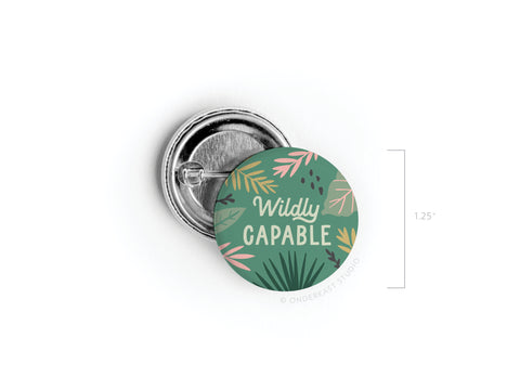 The Onderkast Studio Wildly Capable Pinback Button features vibrant colors on a green background with pink, yellow, and dark green leaves. “Wildly CAPABLE” appears in white and cream text. High-quality; measures 1.25 diameter.