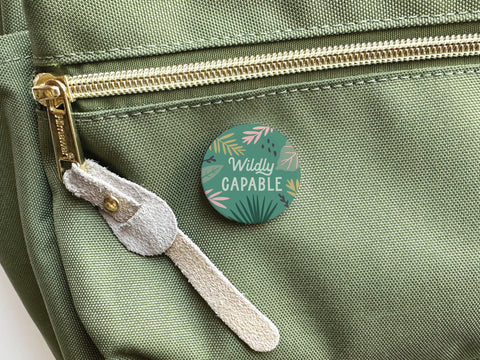 A green fabric bag with a gold zipper and suede pull tab features the Wildly Capable Pinback Button by Onderkast Studio, showcasing a vibrant leafy design in green, pink, and yellow with high-quality printing.