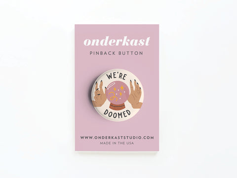 The We’re Doomed Pinback Button by Onderkast Studio features hands around a crystal ball on a lavender card, with onderkast PINBACK BUTTON and www.onderkaststudio.com—crafted for high quality and vibrant detail.