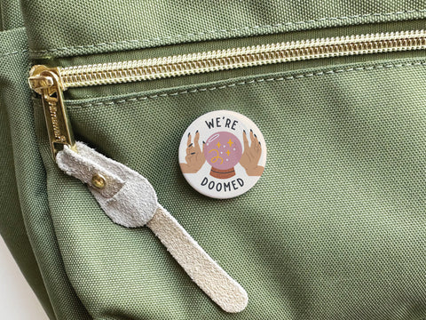 The We’re Doomed Pinback Button by Onderkast Studio features vibrant colors and WERE DOOMED above hands around a crystal ball. It comes on a green fabric bag with a gold zipper pull and suede tab.