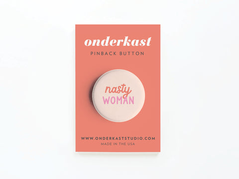The Nasty Woman Pinback Button by Onderkast Studio features vibrant pinks and comes on a coral card labeled onderkast PINBACK BUTTON, with website and Made in the USA at the bottom—a bold accessory for assertive women.