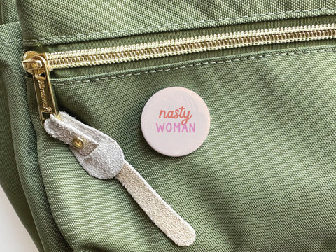 The Onderkast Studio Nasty Woman Pinback Button, featuring bold red and pink lettering, sits atop a green canvas bag with a zippered pocket and suede pull—an ideal accessory for assertive women.