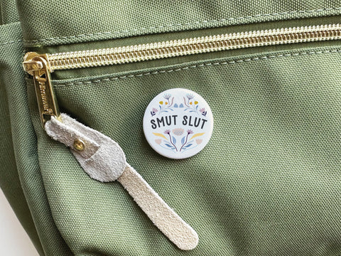 The Smut Slut Pinback Button by Onderkast Studio, featuring floral designs, is attached to the front pocket of a light green bag with a gold zipper and suede pull, adding literary humor to your style.