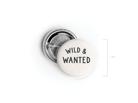 The Wild & Wanted Pinback Button by Onderkast Studio features bold black lettering on white. This round adventure accessory is shown with a 1.25-inch vertical measurement for size reference.
