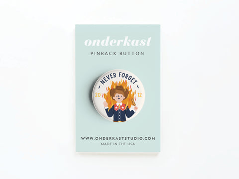 The Never Forget Pinback Button by Onderkast Studio displays a cartoon dog in a suit amid flames with “Never Forget 2012” above, mounted on a mint green card featuring onderkast PINBACK BUTTON, the website, and MADE IN THE USA.