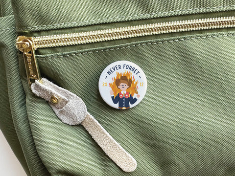 A whimsical green fabric bag with a gold zipper features Onderkast Studio’s Never Forget Pinback Button—a cartoon dog in a suit amid flames, marked with Never Forget, 2012–2022, adding quirky Texan charm.