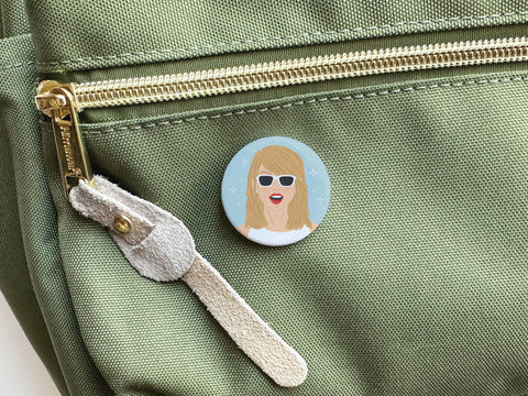 The Onderkast Studio TS 1989 Pinback Button, with artwork of a blonde woman in red lipstick and white sunglasses on blue, is attached to the zipper pocket of a green fabric bag—a subtle tribute to Taylors chart-topping album.