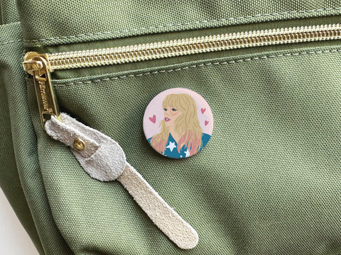 The TS Lover Pinback Button by Onderkast Studio, featuring an illustrated blonde woman and pink hearts, is attached to a green bag near a gold zipper and suede pull, charmingly evoking Taylor Swifts enchanting lyrics.