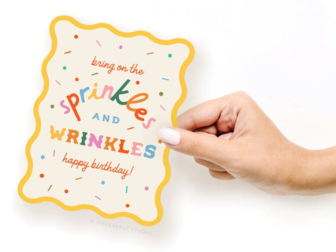 A hand with white nail polish holds the Sprinkles and Wrinkles Wavy Die Cut Notecard by Onderkast Studio, featuring colorful confetti and the phrase “bring on the sprinkles AND WRINKLES happy birthday!” in playful, multicolored fonts.