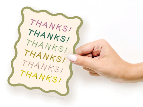 A hand with painted nails holds the Thanks Wavy Die Cut Notecard by Onderkast Studio, featuring “THANKS!” six times in various colors and fonts along its wavy-edged border.