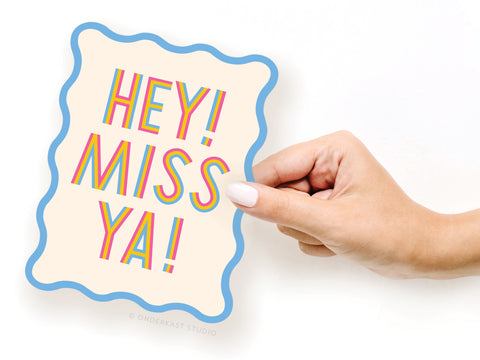 A hand with white nail polish holds the Onderkast Studio Miss Ya Wavy Die Cut Notecard, featuring blue borders and a bold, colorful HEY! MISS YA! design.