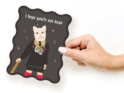 A hand holds the Onderkast Studio TS Reputation Wavy Die Cut Notecard, featuring a cartoon in a cat mask, black outfit, tiger & flowers. i hope youre not dead appears above. The wavy-edged design nods to Taylor Swifts Reputation era.