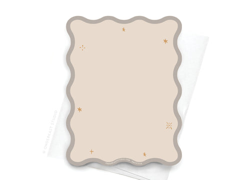 The TS Folklore Wavy Die Cut Notecard by Onderkast Studio in beige features a gray wavy border and small gold star accents, shown placed on a white folded napkin or paper.