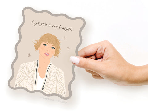 A hand with white nail polish holds the Onderkast Studio TS Folklore Wavy Die Cut Notecard, featuring an illustrated woman with short, wavy hair and a patterned sweater. The card reads, I got you a card—again, on a white background.