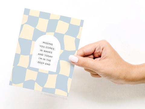 A hand with white nail polish holds the Onderkast Studio Missing You Comes In Waves Greeting Card, featuring a blue and beige checkered pattern and the message: Missing You Comes In Waves and today I’m in the deep end.