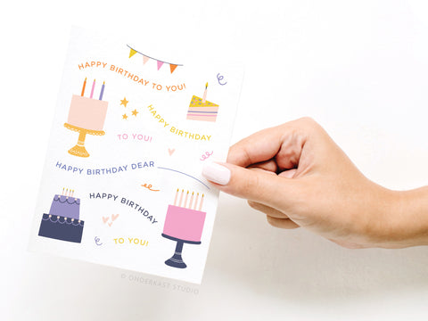 A hand holds the Onderkast Studio Happy Birthday Song Greeting Card, decorated with colorful cakes, banners, and confetti. Featuring playful fonts on premium card stock, it’s perfect for a festive celebration.