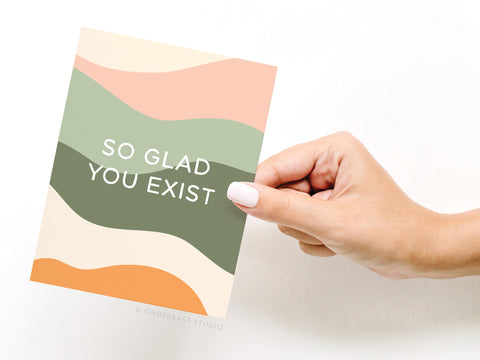 A hand with white nail polish holds the Onderkast Studio So Glad You Exist Greeting Card, featuring abstract pastel wavy patterns and the message SO GLAD YOU EXIST.